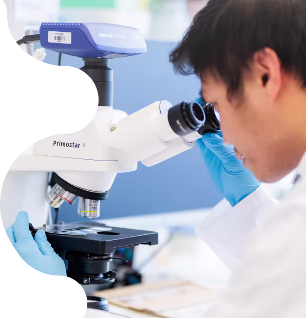 An image of a male scientist wearing a white lab coat and blue gloves. He is looking down a white Primostar 3 microscope.
