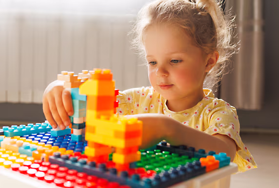 Female toddler with a messy ponytail and yellow t-shirt sits at a table and looks intently at her colourful building block creation, showcasing focus and creativity.